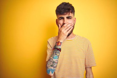 İzole edilmiş sarı arka planda duran çizgili tişörtlü genç adam ağzıyla esnemekten sıkılmış. Huzursuz ve uykulu..