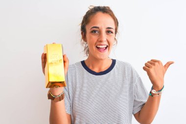 Güzel, kızıl saçlı, altın külçeli genç bir kadın izole edilmiş arka planda, mutlu bir gülümsemeyle baş parmağıyla onu işaret ediyor.