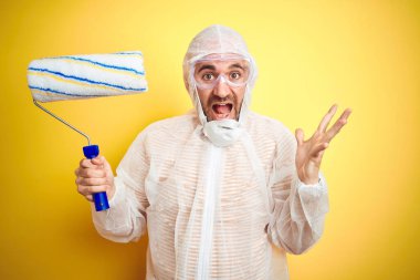 Boyacı kıyafeti giyen ve sarı arka planda boyacı tutan genç adam çok mutlu ve heyecanlı, kazanan yüz ifadesi zaferi büyük bir gülümsemeyle kutluyor ve ellerini kaldırıyor.