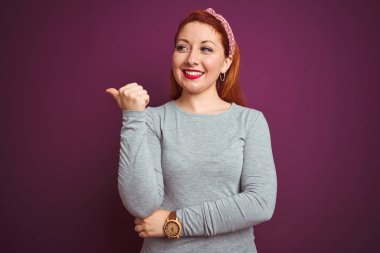 Gri tişört giyen güzel kızıl saçlı kadın ve yalıtılmış mor arkaplan üzerine tebessüm takmış mutlu yüzle gülümseyen ve baş parmağıyla yan tarafı gösteren..
