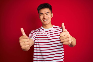 Asyalı çizgili tişört giyen genç bir Çinli izole edilmiş kırmızı arka planda duruyor. Elle olumlu jest yapıyor, gülümsüyor ve başarı için mutlu. Kazanan jest.
