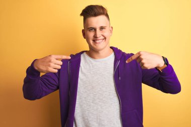 Mor kazak giyen genç yakışıklı adam izole edilmiş sarı arka planda duruyor. Yüzünde bir gülümsemeyle, gururlu ve mutlu bir şekilde kendini gösteriyor..