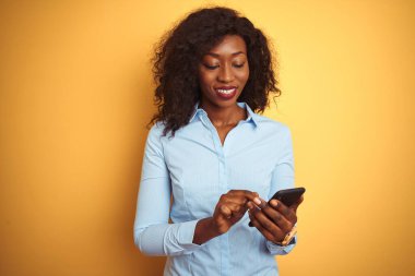 Genç Afro-Amerikalı kadın, izole edilmiş sarı arka planda duran akıllı telefonu kullanıyor. Mutlu bir yüzü var ve kendine güvenen bir gülümsemeyle dişlerini gösteriyor.