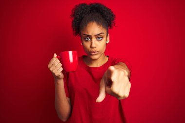 Genç Afrikalı Amerikalı bir kadın izole edilmiş kırmızı arka planda kahve içerken kamerayı işaret ediyor ve sen, el işareti, ön taraftan pozitif ve kendinden emin bir hareket.