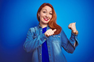 Genç, güzel, kızıl saçlı, kot gömlek giyen, mavi izole edilmiş arka planda duran, el ve baş parmaklarıyla arkasını gösteren, kendinden emin gülümseyen bir kadın.