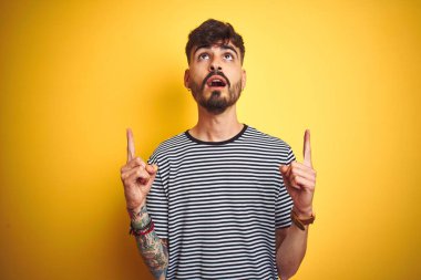 İzole edilmiş sarı arka plan üzerinde duran çizgili t-shirt giyen genç adam şaşırmış ve şaşırmıştı. Parmaklarıyla işaret ediyor ve kollarını kaldırıyordu..