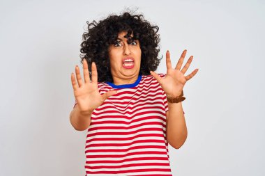 İzole edilmiş beyaz arka plan üzerine çizgili tişört giyen, kıvırcık saçlı genç Arap kadın korku ifadesinden korkuyor ve dehşete düşüyor. El hareketini durdur, şok içinde bağır. Panik konsepti.