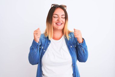 İzole edilmiş beyaz arka planda dikilen kot gömlekli güzel bir kadın. Kolları havada ve gözleri kapalı zaferi kutlarken gülümsüyor. Kazanan konsept.