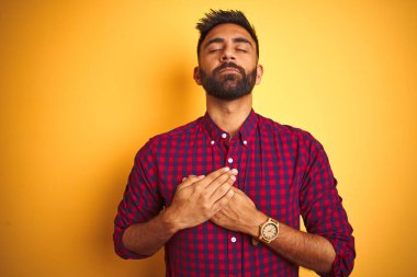 İzole edilmiş sarı arka planda dikilen sıradan bir gömlek giyen genç Hintli adam elleri göğsünde, gözleri kapalı ve yüzünde minnet dolu bir jest ile gülümsüyor. Sağlık kavramı.