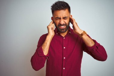 İzole edilmiş gri arka planda duran kırmızı şık gömlek giyen genç Hintli adam yüksek sesli müziğin gürültüsü için parmaklarıyla kulaklarını kapatıyor. Sağır kavramı.
