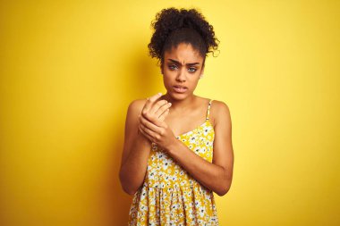 Afro-Amerikalı bir kadın günlük çiçekli bir elbise giyiyor. Sarı arka planda duruyor. Ellerinde ve parmaklarında ağrılar var.