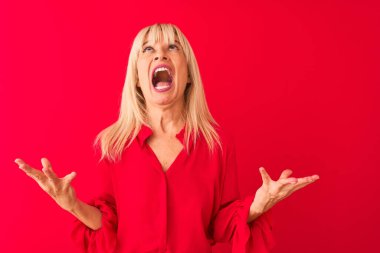 Soyutlanmış kırmızı arka plan üzerinde duran zarif bir gömlek giyen orta yaşlı bir kadın çılgın ve kızgın bir şekilde bağırıyor ve agresif bir ifade ve kollarla bağırıyor. Hayal kırıklığı konsepti.