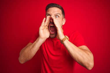 Kırmızı izole edilmiş arka plan üzerine gündelik tişört giyen genç yakışıklı adam elleri ağzında sinirli bir şekilde bağırıyor.
