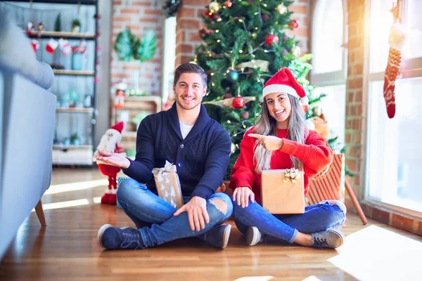クリスマスツリーの周りの床に座っているサンタの帽子をかぶっている若いカップルは 手で提示し 指で指している間にカメラに驚いて笑顔 — ストック写真