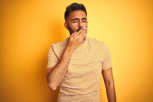 Young indian man wearing t-shirt standing over isolated yellow background bored yawning tired covering mouth with hand. Restless and sleepiness.
