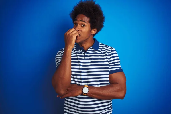 African american man with afro hair wearing striped polo standing over isolated blue background looking stressed and nervous with hands on mouth biting nails. Anxiety problem.