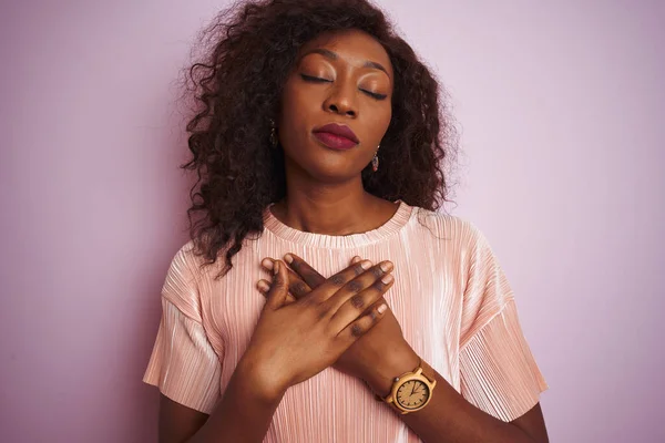 Young african american woman wearing t-shirt standing over isolated pink background smiling with hands on chest with closed eyes and grateful gesture on face. Health concept.