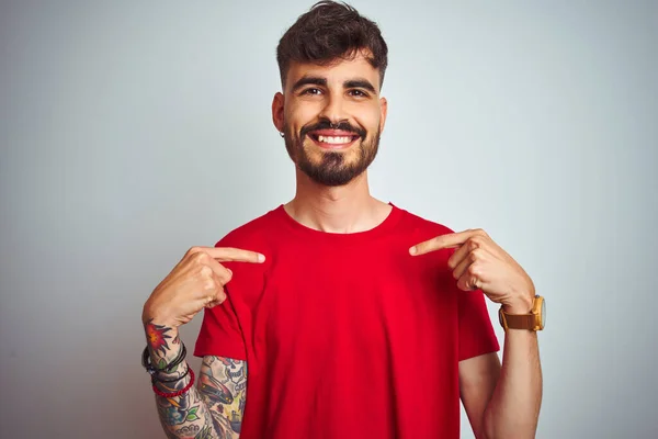 Young man with tattoo wearing red t-shirt standing over isolated white background looking confident with smile on face, pointing oneself with fingers proud and happy.