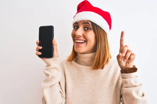 Όμορφη Κοκκινομάλλα Γυναίκα Φορώντας Χριστουγεννιάτικο Καπέλο Δείχνει Οθόνη Smartphone Έκπληκτος — Φωτογραφία Αρχείου