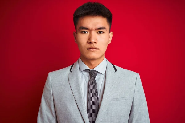亚洲商人身穿灰色夹克 打着领带 站在孤立的红色背景上 脸上带着严肃的表情 简单而自然地看着相机 — 图库照片