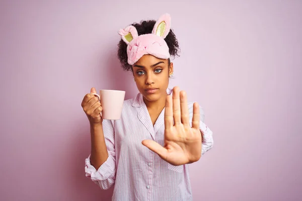 身穿睡衣 戴着面具的非洲妇女 带着严肃而自信的表情和防守姿态 张开手在孤立的粉色背景下喝着一杯咖啡 — 图库照片