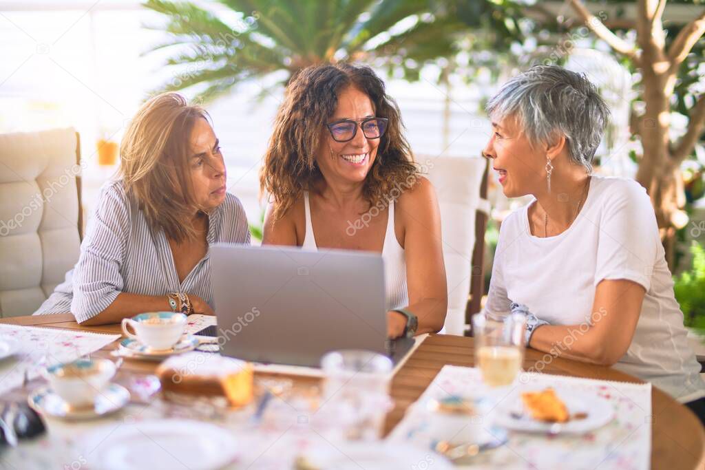 Meeting of middle age women having lunch and drinking coffee. Mature friends smiling happy using laptop at home on a sunny day