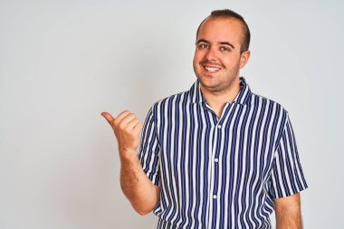 Mavi çizgili tişört giyen genç adam izole edilmiş beyaz arka planda gülümsüyor, gülümsüyor ve başparmağıyla yana bakıyor..