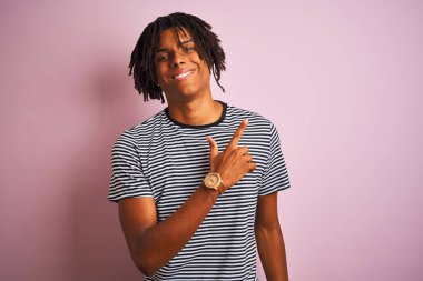 Rasta desenli tişörtlü Afro adam izole edilmiş pembe arka planda mutlu bir şekilde duruyor. Yüzünde mutlu ve doğal bir ifade olan yüzü, eli ve parmağı yana dönük bir gülümseme var.