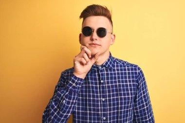 Genç, yakışıklı bir adam, günlük gömlek ve güneş gözlüğü takıyor, izole edilmiş sarı arka planda, elleri çenesinde soru sorarken, düşünceli bir ifade takınıyor. Düşünceli bir yüzle gülümsüyor. Şüphe kavramı.