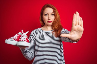 Genç, güzel, kızıl saçlı bir kadın, kırmızı izole edilmiş arka planda spor ayakkabılarını tutuyor. Açık elle dur işareti yapıyor. Ciddi ve kendinden emin bir ifadeyle, savunma jesti yapıyor.