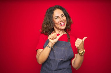 Kırmızı izole edilmiş arka planda önlük giymiş orta yaşlı bir kadın. Arka tarafı işaret ediyor. El ve baş parmakları havada, kendine güvenen.