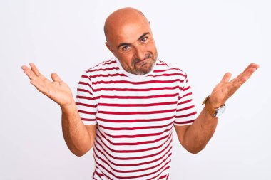 Orta yaşlı, çizgili tişört giyen yakışıklı bir adam soyutlanmış beyaz arka plan üzerinde duruyor. Kolları ve elleri havada, kafası karışmış bir yüz ifadesi var. Şüphe kavramı.