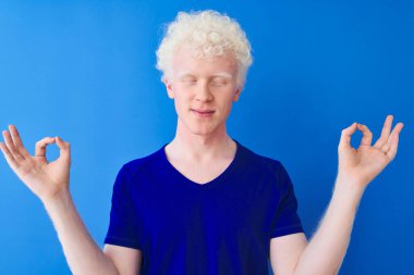 İzole edilmiş mavi arka planda duran sıradan tişört giyen genç albino sarışın adam rahatla ve gözleri kapalı meditasyon yaparken parmaklarınla gülümse. Yoga konsepti.