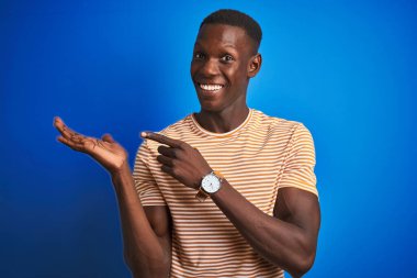 İzole edilmiş mavi arka plan üzerinde duran çizgili günlük tişört giyen Afrikalı Amerikalı adam kameraya hayret ediyor ve gülümsüyor..