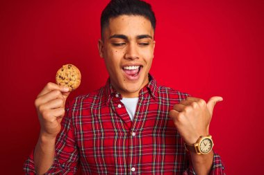 Genç Brezilyalı bir adam elinde kurabiyeyle izole edilmiş kırmızı arka planda duruyor ve gülümseyen bir yüzle baş parmağıyla onu gösteriyor.