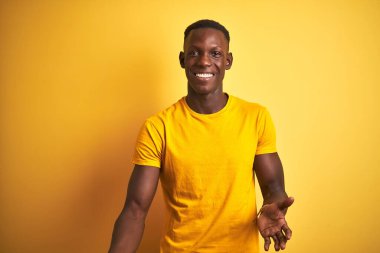 Genç Afro-Amerikan erkeği, günlük tişört giymiş, sarı arka planda dikiliyor, gülümsüyor, kollarını açıp gülümsüyor, dostça karşılıyor, olumlu ve kendinden emin bir şekilde selamlıyor.