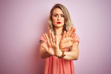 Pembe izole edilmiş arka plan üzerinde duran genç ve güzel bir kadın. Reddedilmiş yüz ifadesi. Kollar ve avuçlar kesişiyor.
