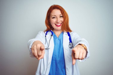 Genç kızıl saçlı kadın steteskop kullanıyor. İzole edilmiş beyaz arka plan üzerinde. Sana ve kameraya parmaklarıyla bakıyor.