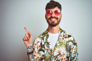 Tatilde dövmeli bir adam, güneş gözlüğü takıyor, izole edilmiş beyaz arka planda, yüzünde kocaman bir gülümseme, eli ve parmağı kameraya bakıyor..