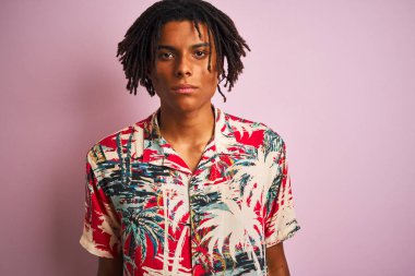 Tatilde saçı rastalı Afro bir adam çiçekli gömlek giyiyor ve pembe bir arka planı var. Yüzünde ciddi bir ifade var. Basit ve doğal bir şekilde kameraya bakıyorsun..