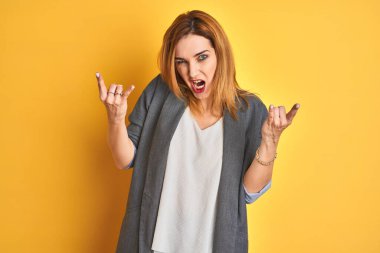 Kızıl saçlı beyaz iş kadını, sarı arka planda izole edilmiş çılgın bir ifadeyle elleri havada rock sembolü yaparken bağırıyor. Müzik yıldızı. Ağır müzik konsepti.