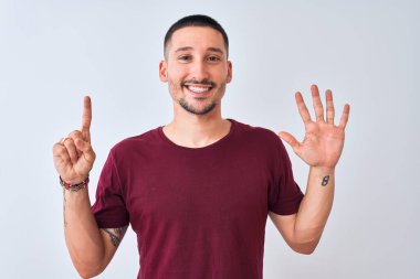 Genç yakışıklı adam izole edilmiş arka planda duruyor ve altı numaralı parmaklarıyla işaret ederken kendinden emin ve mutlu bir şekilde gülümsüyor..