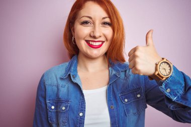 Güzel, kızıl saçlı, kot gömlekli bir kadın pembe arka planda dikilip mutlu bir el hareketi yapıyor. Kameraya bakıp başarıyı gösteren ifade onaylanıyor.