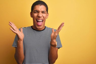 İzole edilmiş sarı arka plan üzerinde donanma çizgili tişört giyen genç yakışıklı Arap adam kollarını kaldırmış ve gözleri kapalı heyecanla haykırarak başarının çılgınlığını kutluyor. Kazanan konsept