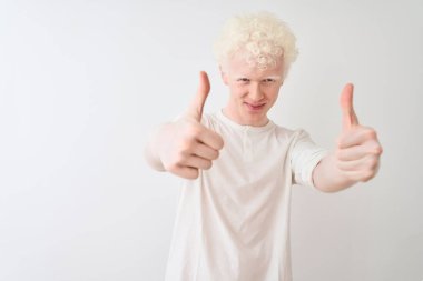 İzole edilmiş beyaz arka planda dikilen sıradan tişörtler giyen genç albino sarışın adam elle olumlu jest yapıyor, gülümsüyor ve başarı için mutlu. Kazanan jest.