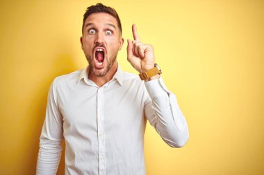 Sarı arka plan üzerine zarif beyaz tişört giyen yakışıklı genç adam başarılı bir fikirle parmağı işaret ediyor. Heyecanlı ve mutlu. Bir numara..