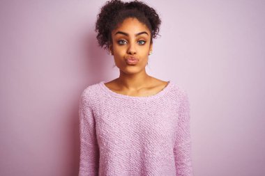 Kışlık kazak giyen genç Afro-Amerikan kadın izole edilmiş pembe arka planda dikilip güzel ve seksi bir şekilde havaya öpücük yollayan kameraya bakıyor. Aşk ifadesi.