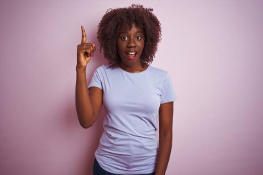 İzole edilmiş pembe arka planda duran ve başarılı bir fikri işaret eden tişört giyen Afrikalı genç bir kadın. Heyecanlı ve mutlu. Bir numara..