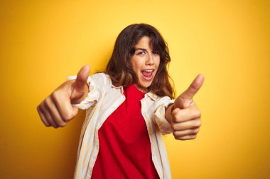 Sarı izole arka plan üzerine kırmızı tişört ve çizgili tişört giyen genç güzel bir kadın el hareketlerini olumlu karşılıyor, gülümseyerek ve başarı için mutlu. Kazanan jest.