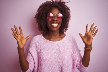 İzole edilmiş pembe arka planda kazak ve güneş gözlüğü takan Afrikalı genç bir kadın başarının çılgınlığını kollarını kaldırmış ve gözlerini kapatmış heyecanla haykırarak kutluyor. Kazanan konsept